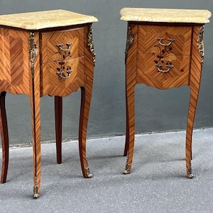 May include: Two antique wooden nightstands with marble tops. The nightstands feature intricate marquetry patterns, floral inlay, and ornate brass hardware. The curved legs and decorative accents add to the classic, elegant design. Each nightstand has two drawers.