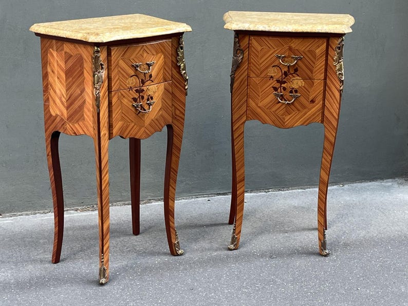 May include: Two antique wooden nightstands with marble tops. The nightstands feature intricate marquetry patterns, floral inlay, and ornate brass hardware. The curved legs and decorative accents add to the classic, elegant design. Each nightstand has two drawers.