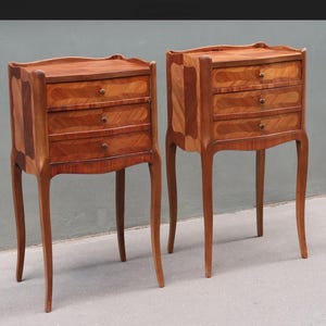 Puede incluir: Dos mesitas de noche antiguas de madera. Cada mesita tiene tres cajones con pomos de latón y patas curvas. La veta de la madera es visible, con tonos marrones claros y oscuros. Las mesitas tienen un diseño clásico y elegante.