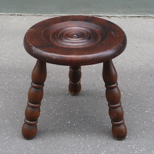 May include: A dark brown wooden stool featuring a circular seat with a carved concentric ring pattern in the centre. The stool is supported by three turned legs, each with a series of rounded, bulbous sections along its length.