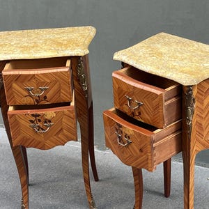 May include: Two antique wooden nightstands with marble tops. The nightstands have two drawers each, with floral inlay details and brass hardware. The wood has a chevron pattern. The legs are curved and the nightstands are in a classic style.