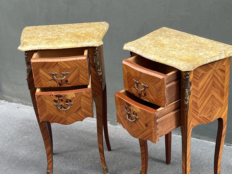 May include: Two antique wooden nightstands with marble tops. The nightstands have two drawers each, with floral inlay details and brass hardware. The wood has a chevron pattern. The legs are curved and the nightstands are in a classic style.