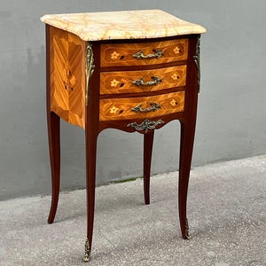 May include: A small antique wooden bedside table with three drawers and a marble top. The drawers are inlaid with floral patterns and the legs are curved.