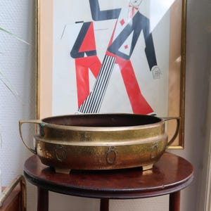 Puede incluir: Una jardinera de latón martillado con dos asas y adornos ovalados, sobre una pequeña mesa redonda de madera oscura. En el fondo, una impresión artística vintage enmarcada con una figura estilizada en rojo, negro y blanco.