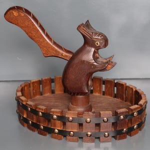 Könnte beinhalten: Ein handgefertigter Nussknacker aus Holz in Form eines Eichhörnchens. Der Nussknacker ist dunkelbraun und steht auf einem runden Holzsockel mit Metallakzenten. Das Eichhörnchen hat ein detailliertes Schnitzdesign.
