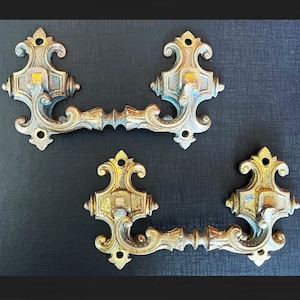 Pode incluir: Duas puxadores de gaveta de metal adornados em tom dourado, com trabalhos em espiral intrincados e um centro decorativo. As puxadores são projetadas para serem montadas horizontalmente.