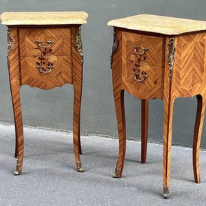 May include: Two antique wooden bedside tables with marble tops. The tables feature intricate inlaid wood patterns, floral drawer details, and brass hardware. Each table has two drawers and curved legs.