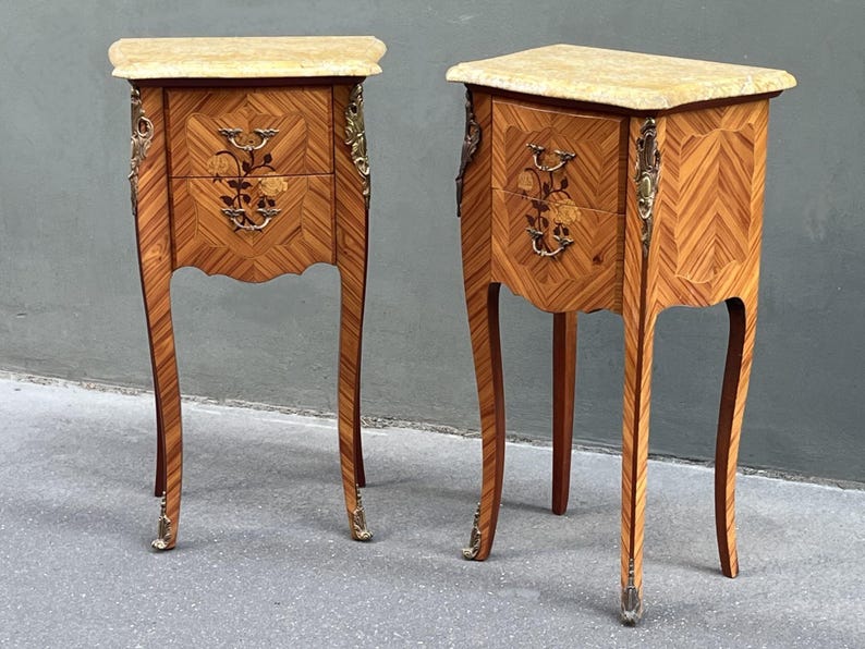 May include: Two antique wooden bedside tables with marble tops. The tables feature intricate inlaid wood patterns, floral drawer details, and brass hardware. Each table has two drawers and curved legs.