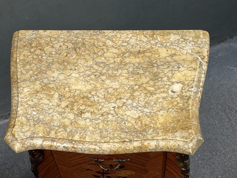 May include: A detailed overhead shot of a small, antique wooden cabinet with a marble top. The marble is a light beige color with intricate brown and gray veining. The cabinet has a decorative drawer pull and ornate carvings.