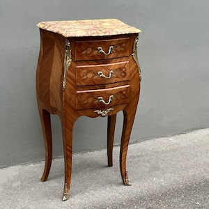 May include: A small, three-drawer bedside table with a marble top. The table is made of wood with a marquetry design and has brass hardware.