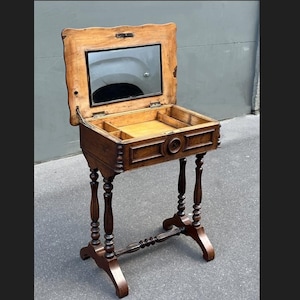 Puede incluir: Una antigua mesa de tocador de madera con una tapa con bisagras que revela un espejo y compartimentos de almacenamiento. La mesa presenta patas torneadas ornamentadas y un travesaño inferior decorativo. La madera tiene un tono marrón cálido.