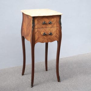 May include: A small, antique wooden nightstand with two drawers. The nightstand has a light-coloured marble top and ornate metal hardware. The wood has a patterned grain and the legs are curved. The nightstand is approximately 71 cm tall.