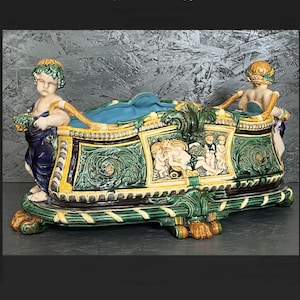 May include: A decorative ceramic centerpiece in a Rococo style. The piece features a rectangular form with a green, yellow, and blue colour scheme. Two cherubic figures flank the sides. The centerpiece has detailed relief carvings and ornate scrollwork.