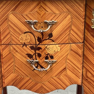 May include: Close-up of a wooden nightstand with a floral inlay design. The nightstand features two drawers with silver handles and a decorative floral motif. The wood has a warm, reddish-brown tone with a chevron pattern.