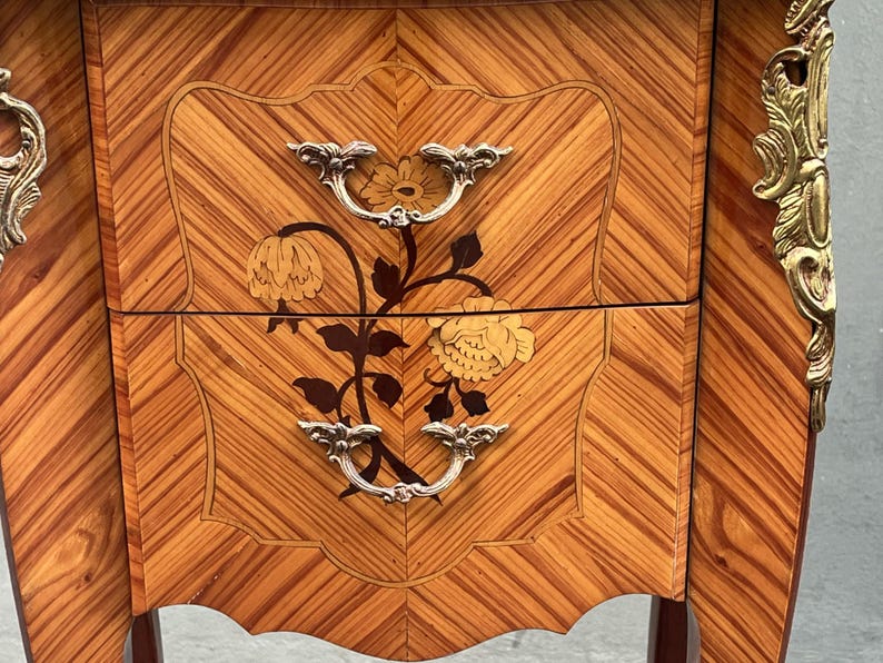 May include: Close-up of a wooden nightstand with a floral inlay design. The nightstand features two drawers with silver handles and a decorative floral motif. The wood has a warm, reddish-brown tone with a chevron pattern.