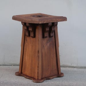 May include: A square wooden pedestal stand with a medium brown finish. The top surface features a circular cutout in the center. The base is flared with a scalloped edge, and the upper section includes decorative corbel-style supports.