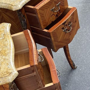 May include: Two antique wooden nightstands with open drawers. The nightstands feature a marble top and ornate metal hardware. The drawers are made of wood with a decorative pattern. The nightstands have curved legs and are set against a gray surface.