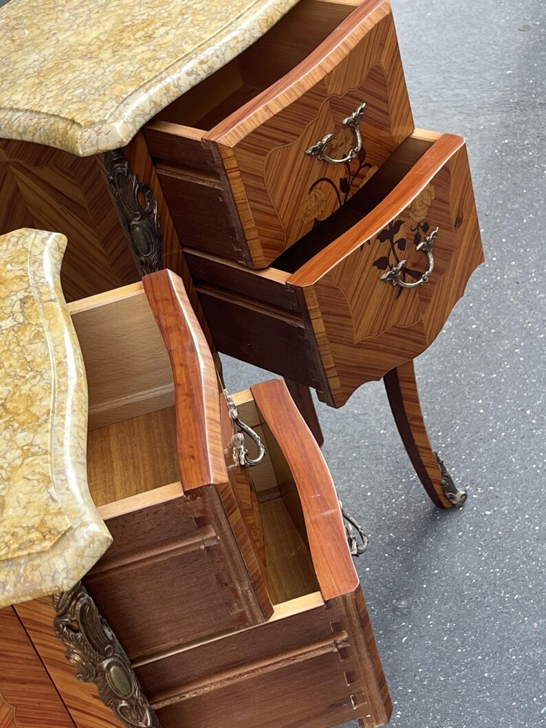 May include: Two antique wooden nightstands with open drawers. The nightstands feature a marble top and ornate metal hardware. The drawers are made of wood with a decorative pattern. The nightstands have curved legs and are set against a gray surface.
