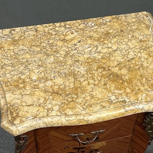 May include: An antique, small wooden cabinet with a marble top. The top is a light beige color with brown veining. The cabinet has a decorative, curved shape and ornate metal hardware. The wood has a patterned design.
