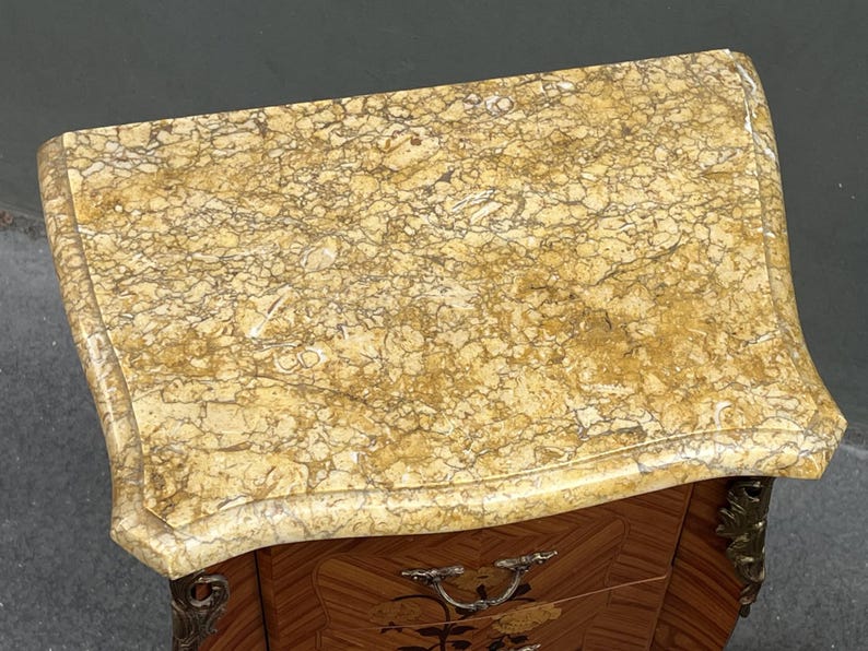 May include: An antique, small wooden cabinet with a marble top. The top is a light beige color with brown veining. The cabinet has a decorative, curved shape and ornate metal hardware. The wood has a patterned design.