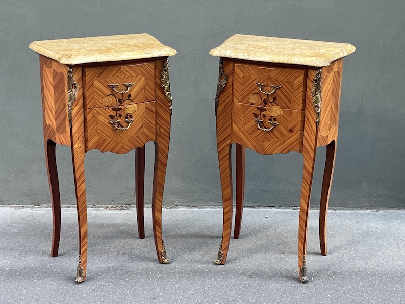 May include: Two antique wooden nightstands with marble tops. The nightstands feature intricate inlaid wood patterns, decorative brass hardware, and curved legs. Each has two drawers with floral accents.