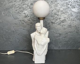 May include: A white ceramic table lamp featuring a sculpture of a couple embracing. The lamp has a round, white globe shade and a brass-colored base. The lamp is plugged into a wall outlet.