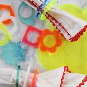 Può includere: Un set di anelli per tovaglioli in acrilico colorati in varie forme, tra cui una stella, un fiore, un quadrato e un esagono. Gli anelli sono su una superficie bianca con un sottopiatto giallo.