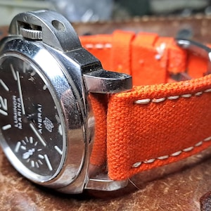 Pode incluir: Um relógio prateado com um mostrador preto e uma pulseira de lona laranja. O mostrador do relógio tem marcas brancas e o texto "Luminor Marina" e "Panerai".
