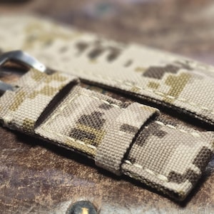 May include: Two beige and brown camouflage patterned watch straps with white stitching. The straps have a silver metal buckle.