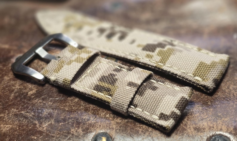 May include: Two beige and brown camouflage patterned watch straps with white stitching. The straps have a silver metal buckle.
