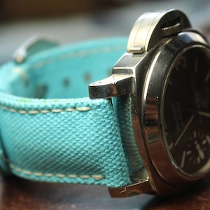 Pode incluir: Close-up de um relógio de pulso com uma bracelete de lona azul-turquesa e um mostrador escuro. O relógio tem uma caixa de aço inoxidável e costuras creme na bracelete. O mostrador exibe os números 6 e 9.