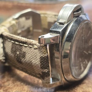 May include: A silver watch with a brown and white patterned canvas strap. The watch has a round face with black hands and markers.