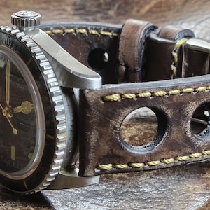 May include: A vintage-style wristwatch with a dark face and silver bezel. The watch has gold hands and markers, and a brown leather strap with decorative holes and yellow stitching. The watch is a classic accessory.