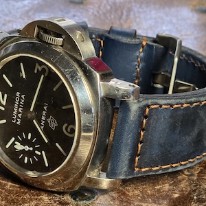 Pode incluir: Um relógio de pulso com mostrador preto, caixa prateada e pulseira de couro azul. O mostrador do relógio tem marcas brancas e o texto "Luminor Marina" e "Panerai" impresso nele.