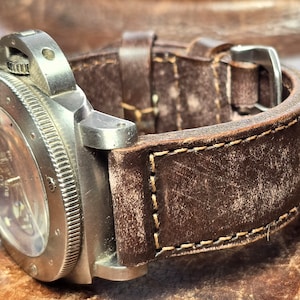 May include: Close-up of a stainless steel wristwatch with a brown leather strap. The watch face is visible, and the strap shows wear. The watch has a textured bezel and a buckle closure. The watch is a luxury item.