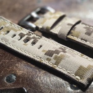 May include: Two beige and brown camouflage patterned watch straps with white stitching. The straps are made of a woven fabric and have a buckle closure.