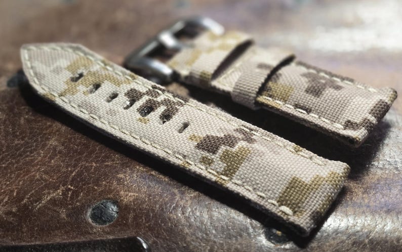 May include: Two beige and brown camouflage patterned watch straps with white stitching. The straps are made of a woven fabric and have a buckle closure.