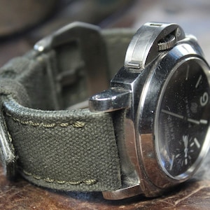 Könnte beinhalten: Eine silberne Uhr mit schwarzem Zifferblatt und einem grünen Canvas-Armband. Die Uhr hat ein Datumsfenster bei 3 Uhr und ein kleines Sekunden-Zifferblatt bei 9 Uhr.