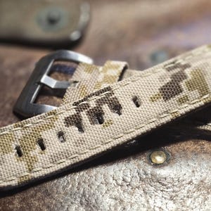 May include: A beige and brown camouflage watch strap with a silver buckle. The strap is made of a woven fabric and has a stitched edge.