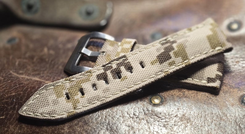 May include: A beige and brown camouflage watch strap with a silver buckle. The strap is made of a woven fabric and has a stitched edge.