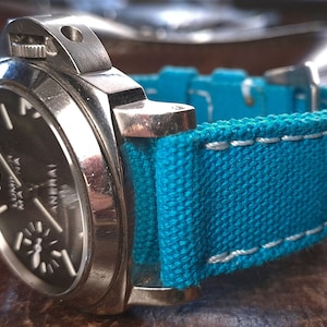 Può includere: Un orologio argentato con quadrante nero e cinturino in tela blu turchese con cuciture bianche.