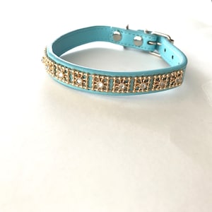 May include: A pale blue pet collar with gold-coloured decorative accents and clear rhinestones. The collar has a silver-coloured buckle and metal hardware. The collar is made of a smooth, leather-like material.