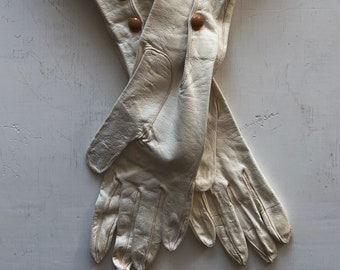 May include: A pair of long, white leather gloves with brown button closures. The gloves are folded in half and the fingers are extended.