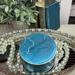 May include: A vintage blue and gold round compact with the text "ELECTRIQUE MAX FACTOR" on the lid. The compact is sitting on a silver tray with white pearl beads.