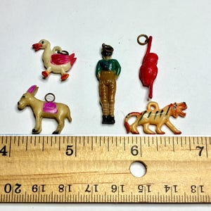 May include: A collection of small, vintage plastic charms. The charms include a duck, a donkey, a man, a monkey, and a tiger. The duck is white and pink, the donkey is tan and pink, the man is wearing a green shirt and brown pants, the monkey is red, and the tiger is tan and orange.