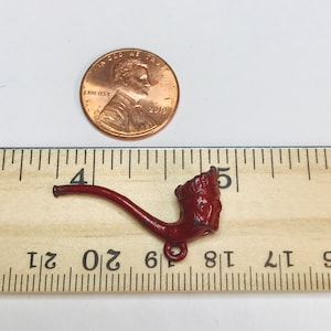 May include: A red metal pipe charm with a detailed design. The charm is about 1.5 inches long and is shown next to a ruler.