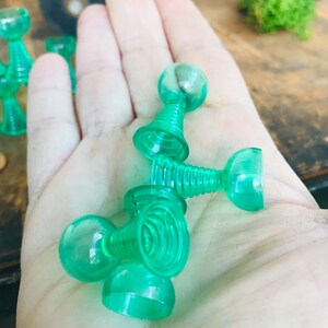 May include: A set of five green plastic toy cups with a ball on top of each cup. The cups are stacked on top of each other.