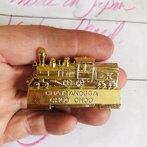 May include: A gold-colored train-shaped pin with the words "Chattanooga Choo Choo" on the front. The pin is held in a hand, and the words "Made in Japan" are visible in the background.