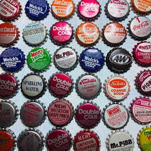 May include: A collection of vintage metal bottle caps featuring various soda brands including Coca-Cola, Double Cola, Sun Crest, Nesbitt's, Welch's, Mr. Pibb, A&W, and Nehi. The caps display colorful logos, brand names, and text on a light background.
