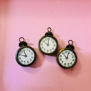 May include: Three small, antique-style clock charms with white faces and black numerals. The clocks have black hands and a bronze-colored metal casing. Each clock shows a different time, and they are arranged on a pink surface.